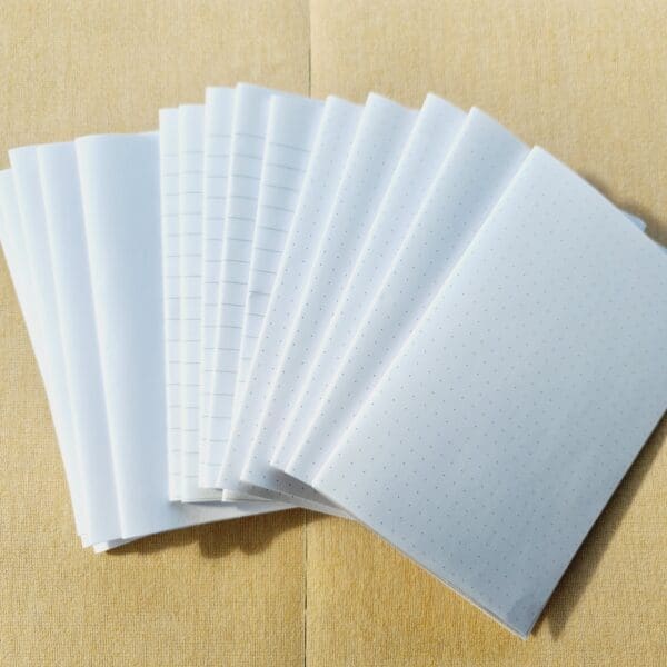 A variety of A6 size paper pages arranged in a fan shape with plain white, ruled lined and dot paper for notebooks by Sarah Lou Crafts.