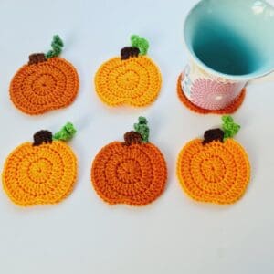 A set of six bright orange, circular crochet pumpkin coasters with a small brown stem and green vines, displayed as a handmade autumn decoration.
