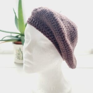 Close-up of a grape coloured crochet beret. The intricate stitch pattern creates a raised, textured surface. The aran yarn is soft and appears thick, ideal for autumn adventures and winter wear.