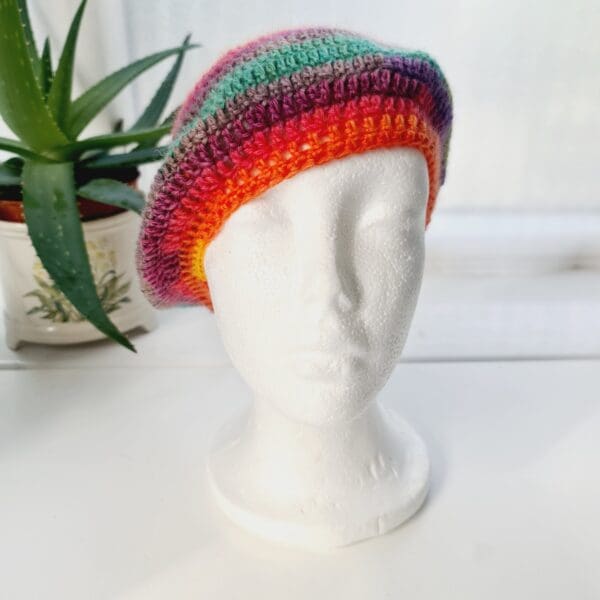 Detailed view of the bright rainbow crochet beret. The edge shows a neat, finished crochet stitch, highlighting the craftsmanship from Sarah Lou Crafts. The texture of the yarn is prominent.