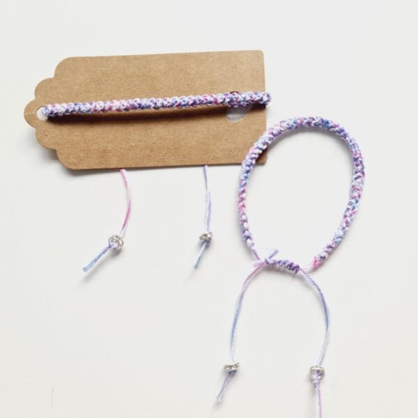 Cord friendship bracelet shown on kraft card with another bracelet next to it without a card, made by Sarah Lou Crafts in the colours of the transgender pride flag.
