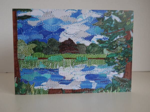 Print of textile collage showing a pond with trees around it and blue sky reflected in it along with clouds.