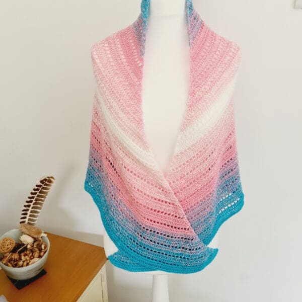 A stylishly draped crochet triangle scarf by Sarah Lou Crafts, showcasing the vibrant Transgender Pride flag colours of blue, pink, and white against a neutral background.