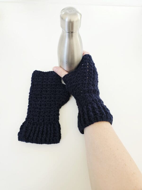 A pair of navy blue crochet fingerless mitts is worn on a person's hands as they hold metal water bottle. This image shows the mitts being worn in a natural, active context, emphasising their functionality for outdoor adventures.