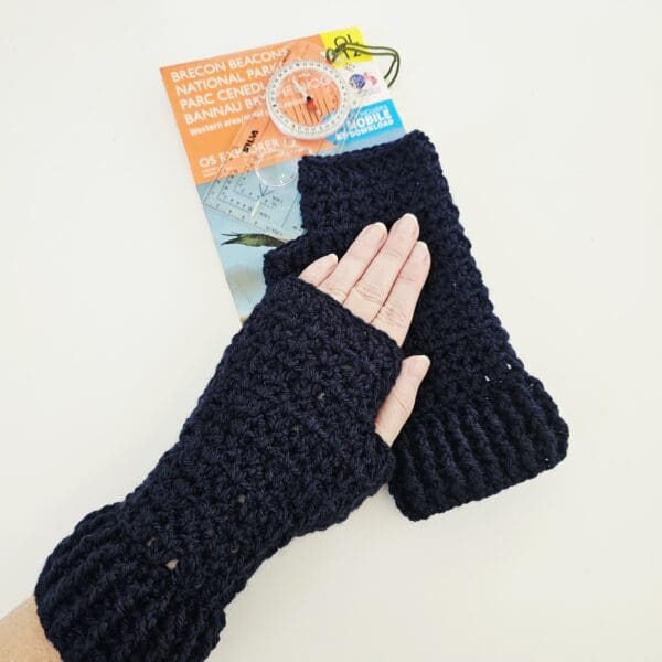A close-up of a person's hand, wearing one navy blue crochet fingerless mitt, laying flat on the other mitts on top of a map and compass. This shot focuses on the mitt and the compass, drawing attention to the detail of both items and reinforcing the outdoor theme.