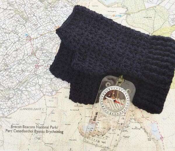 A pair of navy blue crochet fingerless mitts sits on a hiking map, with a compass and nearby. This is a top-down shot showing the mitts and the hiking gear together, suggesting they are a practical accessory for outdoor activities.