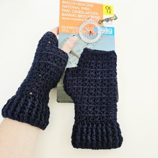 One navy blue crochet fingerless mitt is shown in a close-up, worn on a hand holding a compass, with an OS hiking map in the background. This shot focuses on the texture of the mitt and its use, highlighting the freedom of movement for the fingers while keeping the hand warm.