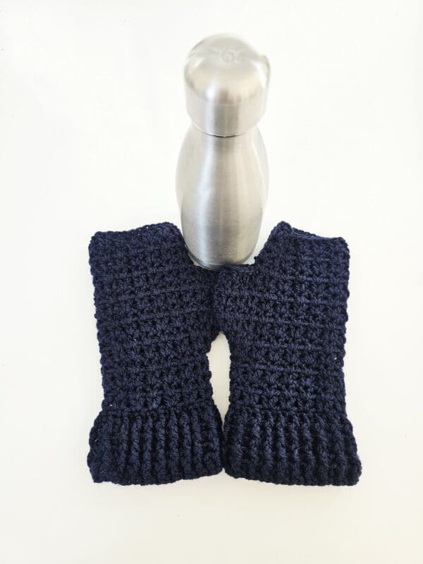 A pair of navy blue crochet fingerless mitts laying flat next to a water bottle. This is an action shot that demonstrates the mitt's practicality and ease of use with other objects.