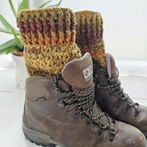 A pair of dark brown hiking boots shown from the side. A pair of crochet boot cuffs is visible showing the autumn colours of burnt orange and brown. The boot cuffs have a ribbed texture.