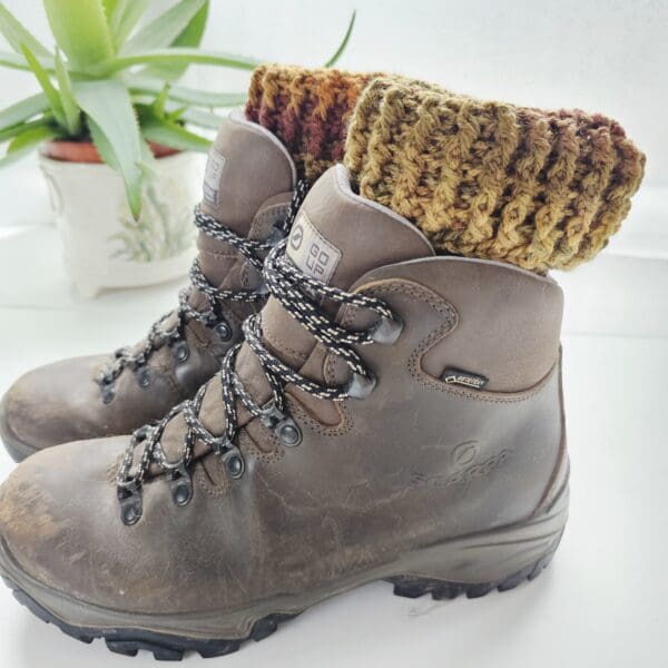 A pair of brown hiking boots with crochet boot cuffs in autumn colours, peeking out from the top. The boot cuffs have a textured, ribbed pattern.