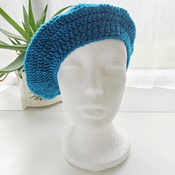 Close-up of a hand-crocheted beret hat on a white mannequin head. The hat is made from Sea Blue aran yarn and features a tightly-worked crown and a neat, ribbed brim.