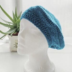 Beautifully crafted Sea Blue aran crochet beret modelled on a stark white display head, showcasing its rich colour, warm texture, and perfect finish.