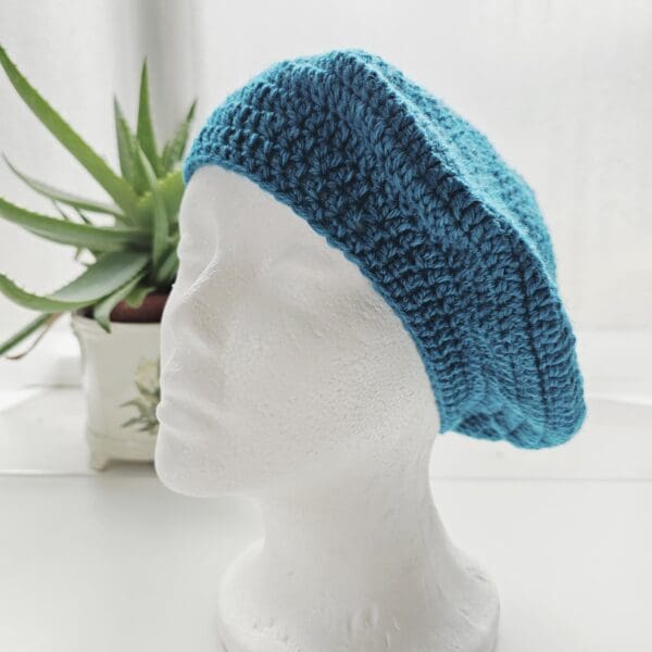 Beautifully crafted Sea Blue aran crochet beret modelled on a stark white display head, showcasing its rich colour, warm texture, and perfect finish.