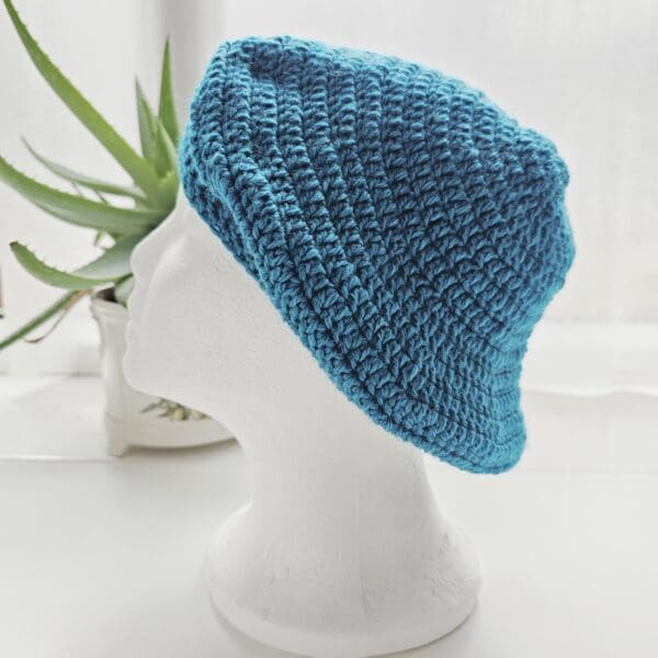 Handmade Sea Blue aran crochet beret displayed on a white mannequin, showing the hat from a left side angles, including close-ups of the stitches and the overall finished shape.