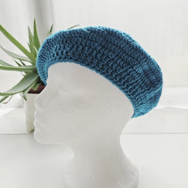 Side profile of a classic crochet beret in Sea Blue on a white mannequin head, highlighting the signature slouchy shape and the tapered band around the base.