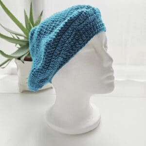 Full view of a stylish crochet beret in a vibrant Sea Blue colour, displayed on a clean, white mannequin. The aran yarn gives the hat excellent texture and drape.