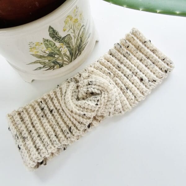 A close-up of a cream and black speckled crochet ear warmer headband on a white mannequin head. The focus is on the wide twist knot at the front, highlighting the texture and detail of the stitches.