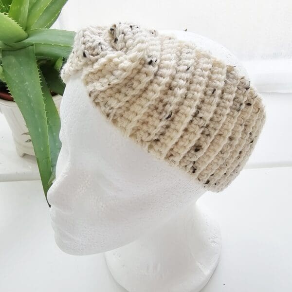 A full-length shot of a white mannequin head wearing the cream and black speckled crochet ear warmer headband. The headband sits just below the hairline and covers the top part of the ears.