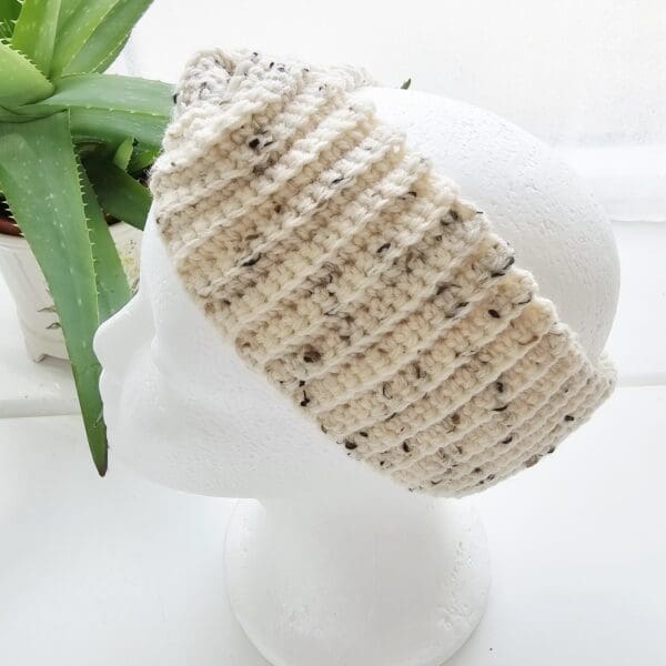 A cream and black speckled crochet ear warmer headband, made from aran yarn, is worn on a white mannequin head. The headband has a wide twist knot at the front. The background is a light, blurred studio setting.