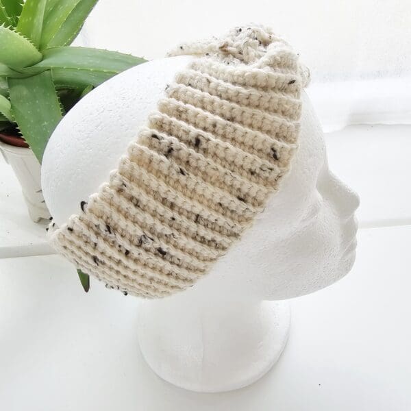 A side view of the cream and black speckled crochet ear warmer headband on a white mannequin head, showing how it fits comfortably over the ears and the width of the band.