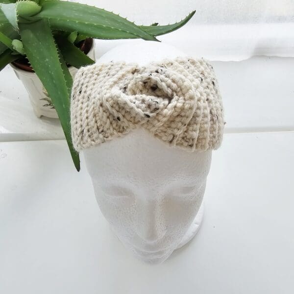 A slightly elevated, top-down view of the cream and black speckled crochet ear warmer headband on a white mannequin head, providing a clear perspective of the headband's shape and the texture of the yarn.