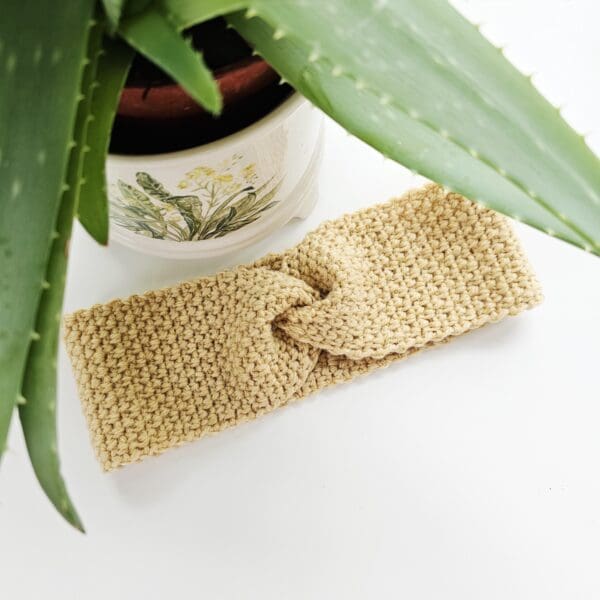 Studio photo of a beige/sand coloured aran yarn crochet ear warmer for cold weather, modelled next to an aloe vera plant.