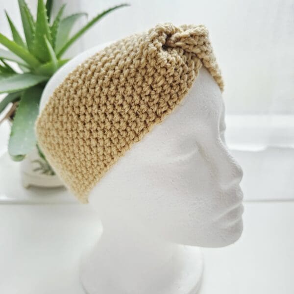 Close-up of a textured crochet ear warmer in a neutral sand colour, modelled on a white head form.