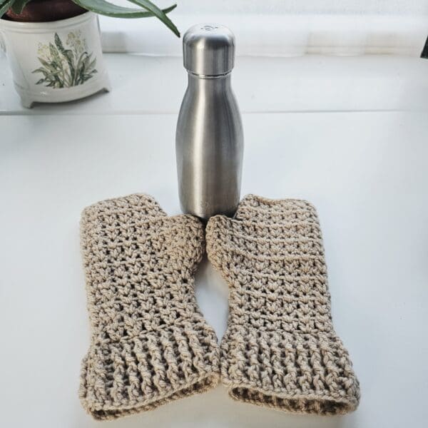 A pair of coffee-coloured crochet fingerless mitts draped over a modern stainless steel water bottle