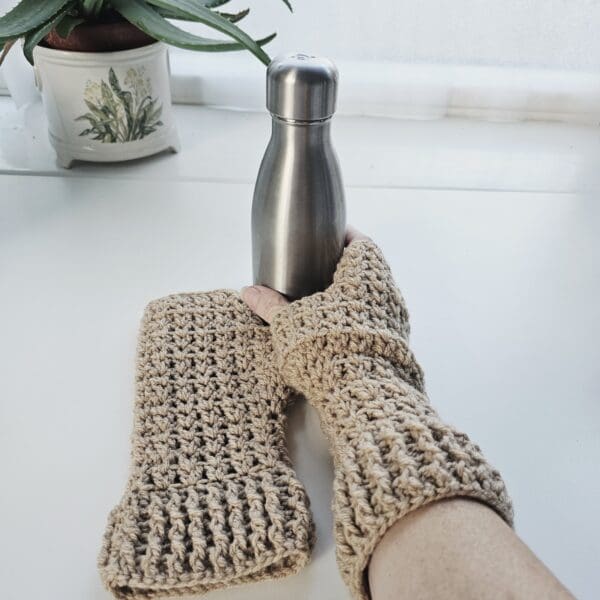 Crochet fingerless mitts in a warm light brown shade, with the cuff detail visible, displayed holding a sleek, insulated water bottle.