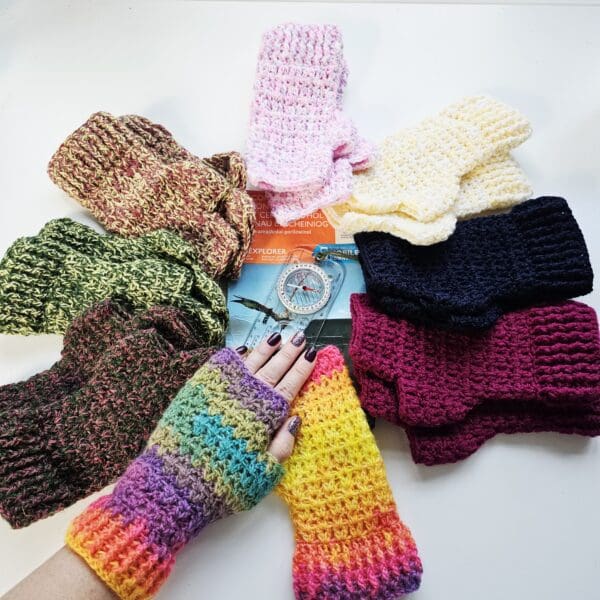 A group of eight pairs of crochet fingerless mitts are arranged around a folded Ordnance Survey map. The mitts, in a variety of colours including rainbow, green, pink, yellow, and variegated yarns, are laid flat, with each pair positioned so the map forms a central point. The mitts and map are presented on a light-coloured surface.