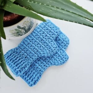 Crochet fingerless gloves in sky blue acrylic yarn, shown on a white table with aloe vera plant to the side.