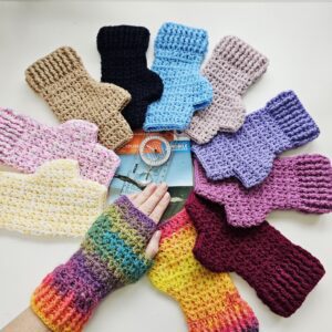 A group of ten pairs of crochet fingerless mitts are arranged around a folded Ordnance Survey map. The mitts, in a variety of colours including rainbow, green, pink, yellow, and variegated yarns, are laid flat, with each pair positioned so the map forms a central point. The mitts and map are presented on a light-coloured surface.