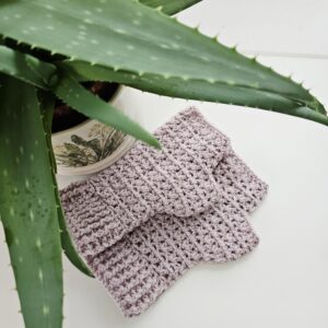 Crochet fingerless gloves in pastel pink acrylic yarn, shown on a white table with aloe vera plant to the side.