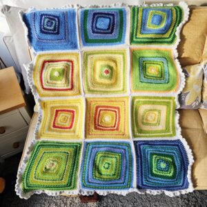 A full-length view of a hand-crocheted temperature blanket laid flat on a double bed. The stripes transition through a spectrum of colours, from icy blues and soft greys to warm ochres and deep oranges, representing a full year of British weather.