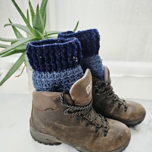 A close-up side profile of a sturdy brown leather hiking boot. Peeking out from the top is a thick, ribbed crochet boot cuff in two-tone navy and royal blue. The texture of the wool is chunky and snug against the ankle.