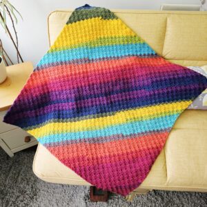 A vibrant, multicoloured crochet blanket draped neatly over the back of a bright yellow fabric sofa. The blanket features a repetitive geometric pattern in shades of teal, mustard, cream, and coral, contrasting boldly against the sunny yellow upholstery.