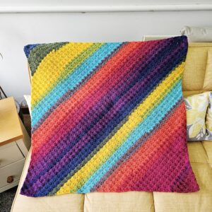 A close-up shot of a handmade multicoloured crochet blanket resting on a yellow sofa. The image highlights the intricate "granny square" stitching and the soft, woolly texture of the yarn. A corner of the blanket hangs over the edge of a yellow cushion.
