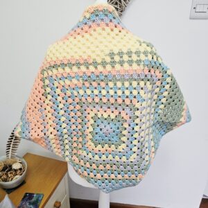 A close up shot of the crochet work, showing the intricate "granny stitch" pattern. The yarn features a soft, matte finish in a variegated pastel colourway including lemon yellow and sky blue.