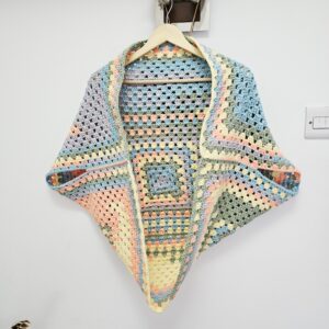 A front-facing view of a handmade crochet cardigan shrug in pastel shades of mint, lilac, and soft peach. It is displayed hanging against a neutral background, showing the open-front design and cropped waistline.