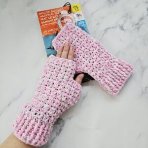 Close-up of a hand wearing a soft pink and white crochet fingerless glove, holding a clear plastic baseplate compass. The soft, feminine colours of the wool contrast with the technical look of the navigation tool.