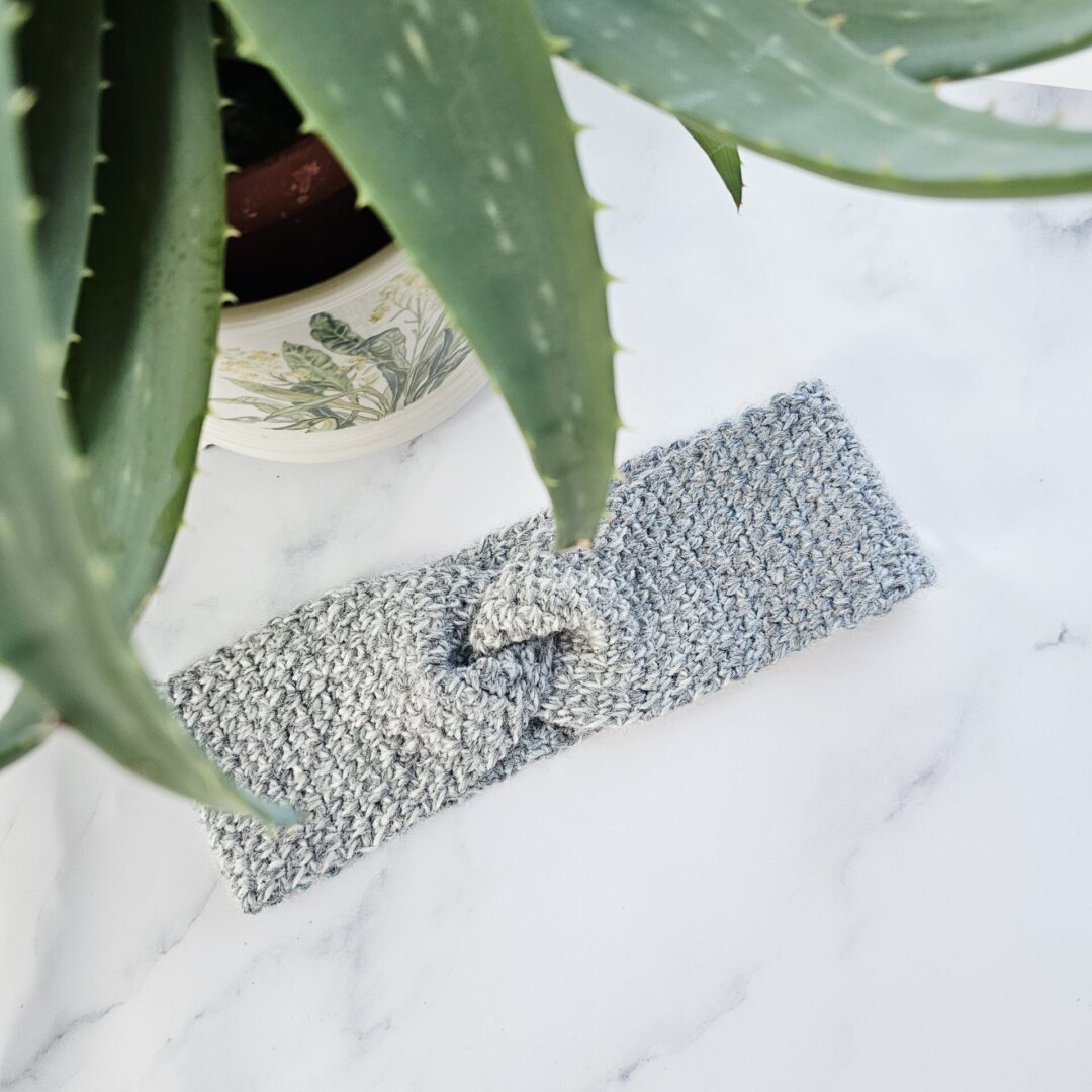 A marble grey crochet ear warmer with a central twist detail, displayed on a mannequin head against a clean, neutral background. The lighting highlights the depth of the grey tones and the comfortable nature of the knit.