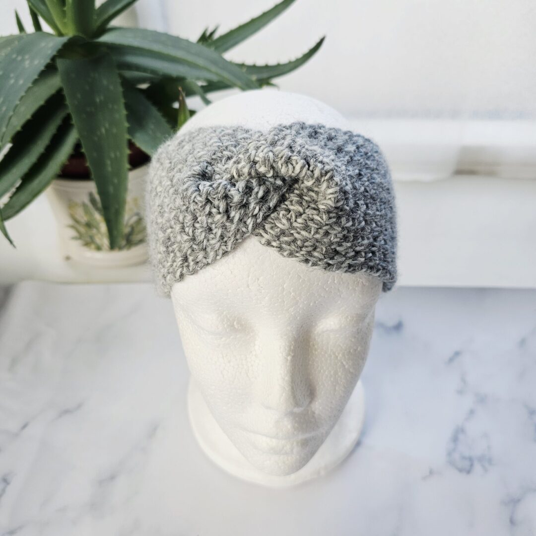 A marble grey crocheted ear warmer shown on a white mannequin head. The headband features a stylish central twist or knot detail at the forehead, showcasing a chunky, textured stitch pattern in a mix of light and dark grey tones.