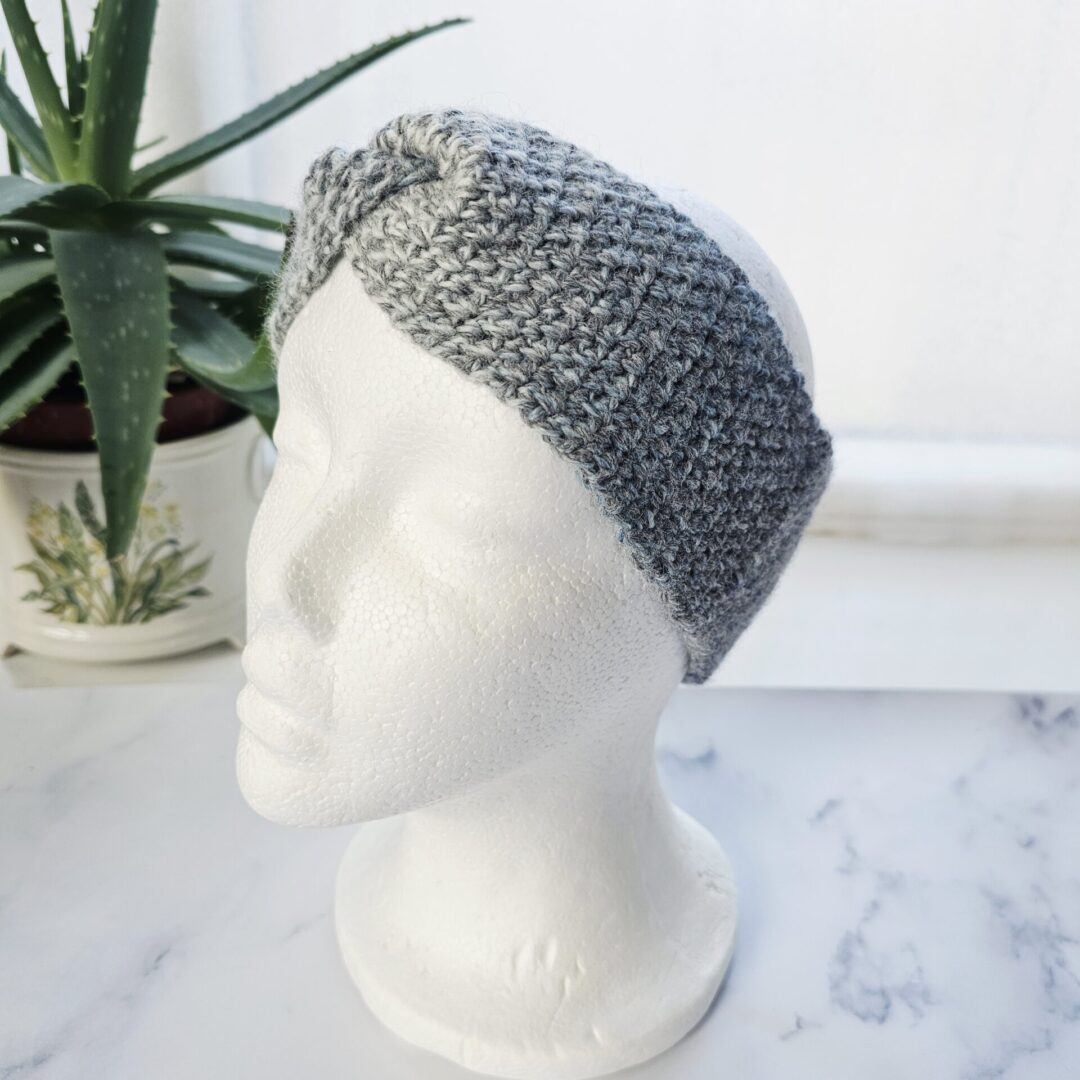 An angled view of a white mannequin head showing the seamless join of a marble grey crochet ear warmer. The band is wide and substantial, designed to sit comfortably under or over hair for winter warmth.