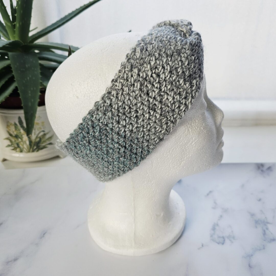Side profile of a mannequin wearing a marble grey crochet headband. The wide design completely covers the ears, highlighting the thick, warm texture of the wool and the intricate ribbed crochet stitches.