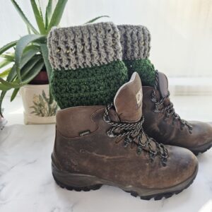 A close-up side profile of a sturdy brown leather hiking boot. Peeking out from the top is a thick, ribbed crochet boot cuff in two-tone green and sage. The texture of the yarn is chunky and snug against the ankle.